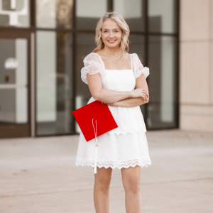 hannah standing with her arms crossed in a white dress holding her crimson red graduation cap standing in front of a building with large glass doors