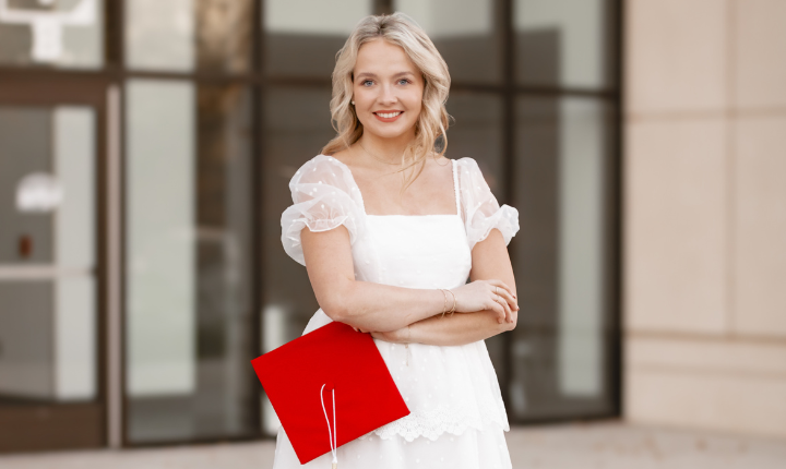 hannah standing with her arms crossed in a white dress holding her crimson red graduation cap standing in front of a building with large glass doors